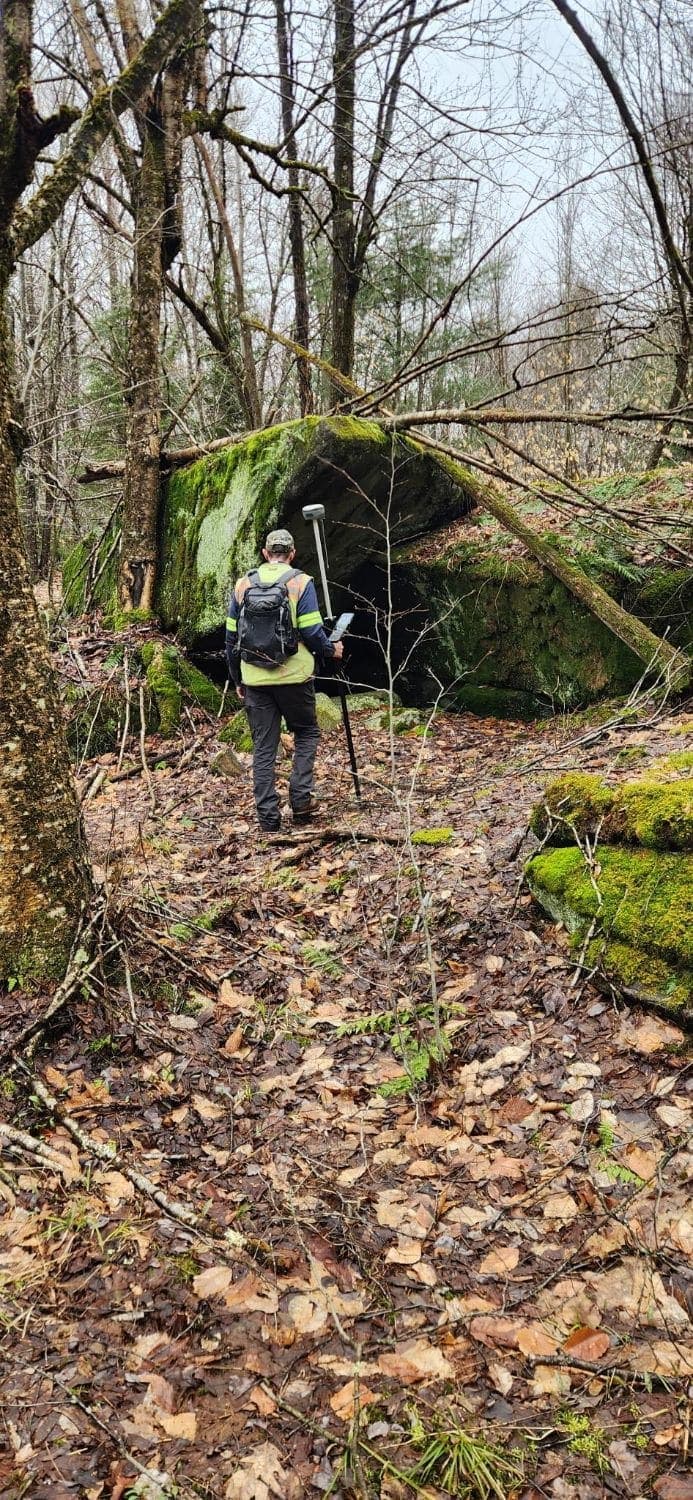 A surveyor with a GPS rover rod and data collector beside a large moss-covered boulder in bare late-autumn woods