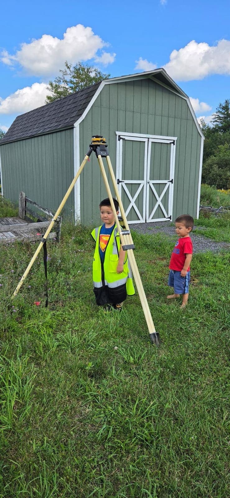 Two small boys beside a survey tripod in a yard, one wearing an oversized safety vest