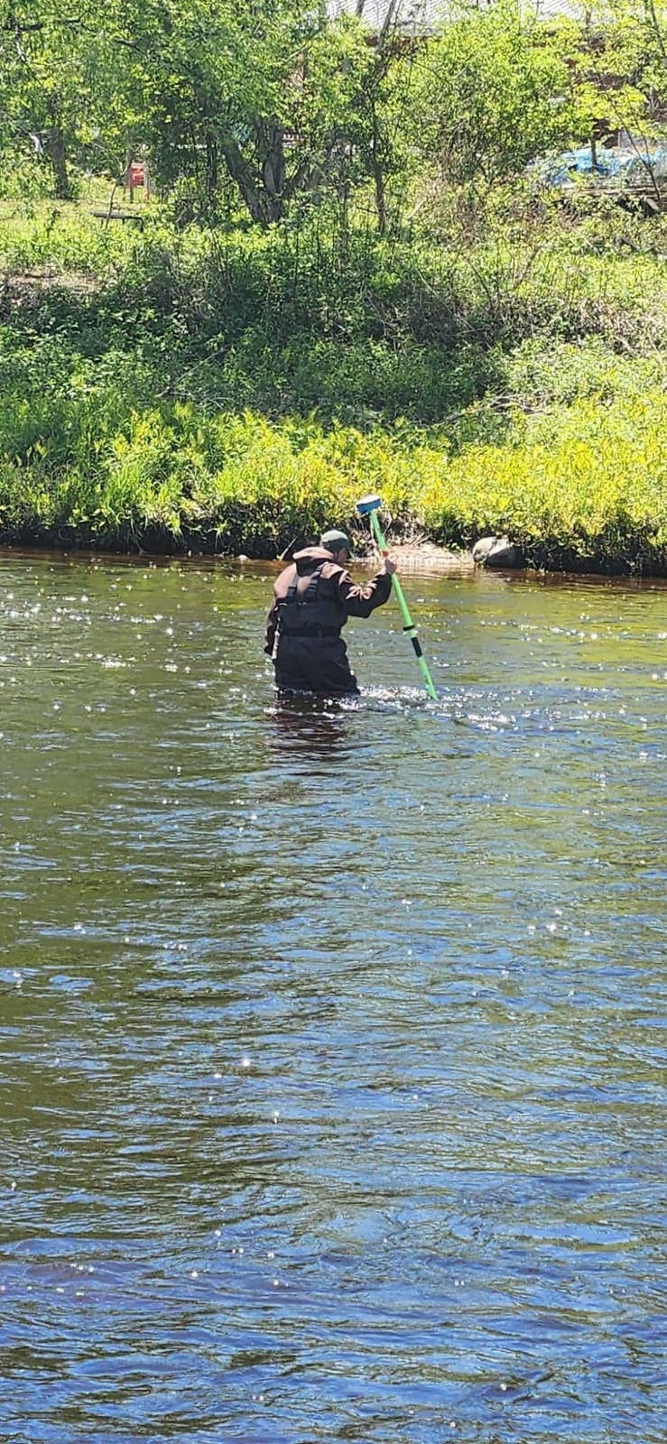 A surveyor in waders wading a river with a GPS rod, green bank behind